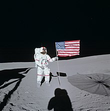 An astronaut in an Apollo space suit with red stripes on the arms and legs and down the helmet stands amid gray dust, grasping the pole of an American flag