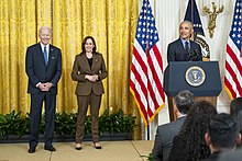 Photo of Obama standing behind a lectern, giving a speech at the White House, with Biden and Harris smiling in the background