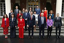 Sanchez wearing a suit and red tie standing with his cabinet surrounding him