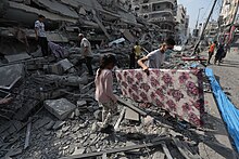 People stand amid the rubble of a building and looking at the ground. A man is carrying a large flower-patterned object.