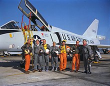 The astronauts pose in front of a delta-winged light blue-gray jet aircraft, holding their flight helmets under their arms. The three Navy aviators wear orange flight suits; the Air Force and Marine ones are green.