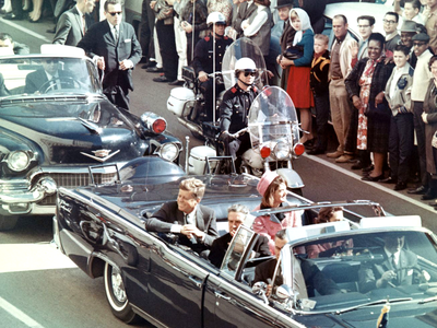 John F. Kennedy and others ride in a black roofless Lincoln Continental convertible down a street lined with spectators, flanked by police officers on motorcycles, and followed by Secret Service officers.