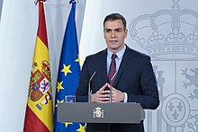 Pedro Sanchez standing behind a podium with the flags of Spain and the EU in the background online with the Spanish crest partially visible on the wall behind him