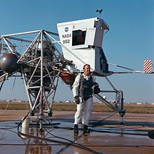 Shepard stands in a white flight suit in front of a vehicle made of tubing, with two metal spheres and a small cabin
