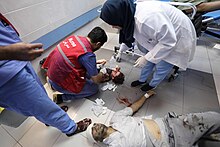 A bloodied infant on the floor of a hospital being treated by a nurse. A man with his head wrapped in a bandage wearing blood-stained clothes lays on the floor nearby.