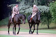 Elizabeth and Ronald Reagan on black horses. He bare-headed; she in a headscarf; both in tweeds, jodhpurs and riding boots.