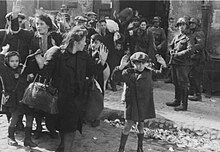 A young boy surrounded by other unarmed civilians holds his hands over his head while a man in uniform points a submachine gun in his direction