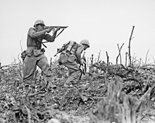 a black and white image of two Marines in their combat uniforms. One Marine is providing cover fire with his M1 Thompson submachinegun as the other with a Browning Automatic Rifle, prepares to break cover to move to a different position. There are bare sticks and rocks on the ground.