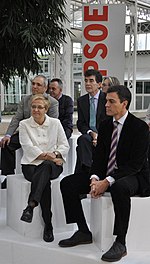 Pedro Sánchez in a suit and tie sitting next to a woman wearing a white top and black pants with several people standing behind them.