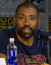 A photograph of Cress Williams speaking at a convention behind a microphone