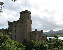 A grey castle with tall square towers stands amongst trees in full leaf