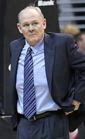 A man with gray hair, wearing a black suit, white shirt and tie, sitting at a basketball game.