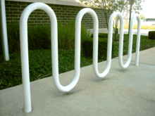A white-painted round metal tube bent into a double M shape and set in concrete in front of a grassy verge and a brick wall.
