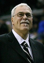 A man with gray hair, wearing a black suit, white shirt and tie, at a basketball game.