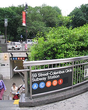 One of the entrances to the 59th Street–Columbus Circle station, located in a sunken plaza with trees inside it. There is a sign on a gray fence next to the entrance. The sign contains the text "59 Street–Columbus Circle Subway Station" and the icons of the "A", "C", "B", "D", and "1" trains.