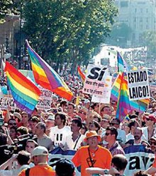 一大群西班牙人在陽光普照的街道上游行, 他們很多人高舉代表同性戀自豪的彩虹條紋旗幟, 也有人舉起標語牌, 其中照片中最為顯眼的一個寫著"ESTADO LAICO", 意為世俗國家, 另一個寫著"Si".
