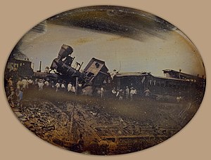 Black and white photograph of a train wreck; one locomotive is on top of the other and several train cars are demolished. A crowd of bystanders surrounds the wreck.