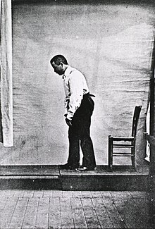 黑白照片中的帕金森氏病患者走路時身體佝僂，照片由他的左邊拍攝，而他的後方有一張椅子。