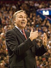 A man with gray hair, wearing a black suit, white shirt and tie, at a basketball game.