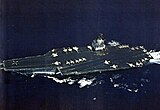 Large aircraft carrier with boxy island superstructure and numerous aircraft on its flight deck.
