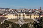 Rococo three- and four-story palace stretches across most of the midground. In the foreground are manacured lawns and walkways, while the background is the old city of Vienna with a cathedral on the horizon.