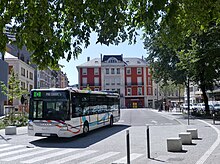 Photographie en couleurs d’un autobus blanc avec des traits multicolore quittant un arrêt de bus en zone urbaine, avec un bâtiment rouge et blanc dans le fond. Invisible sur la photo, la gare ferroviaire se trouve à gauche.