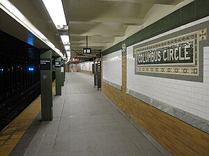Uptown platform of the IRT station. To the left is a set of green columns and a yellow strip at the edge of the platform. To the right is a tiled wall, which contains a mosaic sign with the words "Columbus Circle".