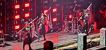 Three women standing on stage carrying flags over their shoulders. The flags contain the Babymetal logo. A drummer is visible in the back, and a guitarist on the very right, cut off by the edge of the image.