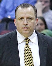 A man with dark hair, wearing a black suit, white shirt and tie, at a basketball game.