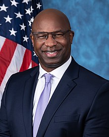 Bowman smiling in a navy blue suit with a purple tie and white shirt, in front of an American flag