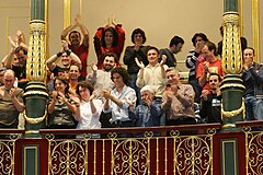 二十多個身穿便裝的成年西班牙男女, 隔在裝飾精美的欄桿和柱子後的旁聽席站起和愉快地拍掌.