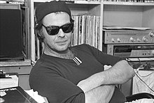 black & white shoulder high portrait of man in his thirties with folded arms seated at desk in front of vinyl record albums and a stereo, wearing black sunglasses, dark backwards baseball cap, and dark shirt