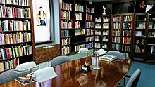 Wooden library shelves and wood study table with lamp. Many books line the shelves along with small human figurines. A sunlit window frames a stained glass outline of a human in leather vest and chaps.