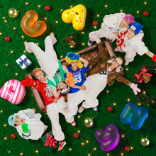 The members are smiling towards the camera while they are standing on a grass field or laying on a couch. They are surrounded by candies of various size and color and other Christmas decorations.