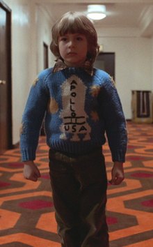 Danny Lloyd as the child character Danny Torrance in the 1980 film The Shining. He stands in a hotel hallway and wears a sweater with a rocketship on it.